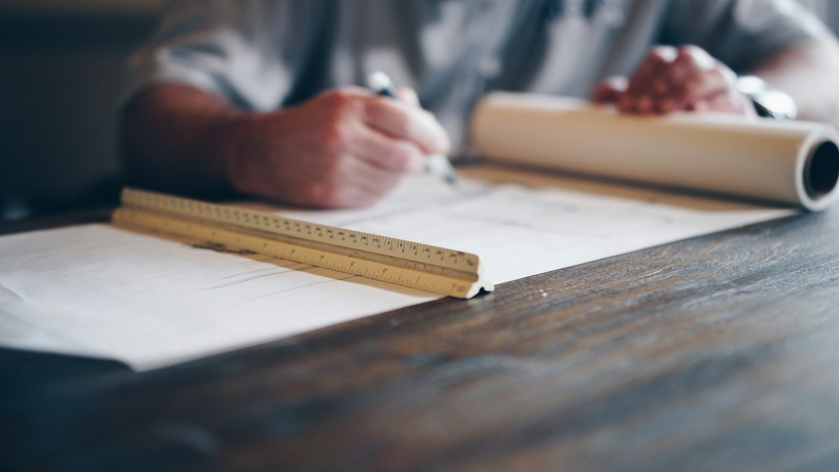 A person at a dark wooden desk drawing on a plan, with a wooden scale rule in the foreground and a rolled drawing alongside.