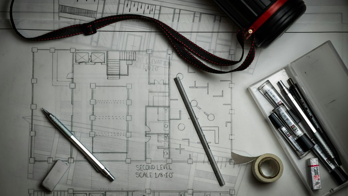 A hand-drafted architectural floor plan laid flat on a desk, with mechanical pencils, an eraser, a metal scale rule and a drawing tube resting on top of it.