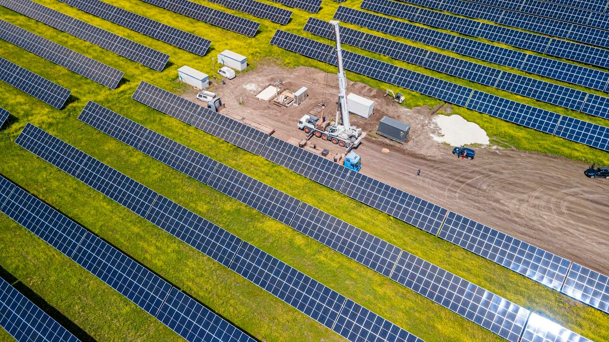 An aerial view of a solar farm under construction, with rows of panels, a mobile crane and a site compound of containers and vehicles