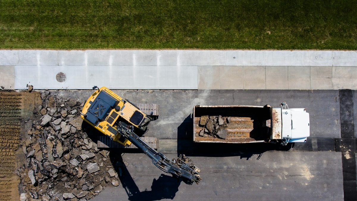 An overhead view of a yellow tracked excavator with a breaker attachment working beside a white dump truck on a stretch of road being torn up, broken asphalt piled alongside.
