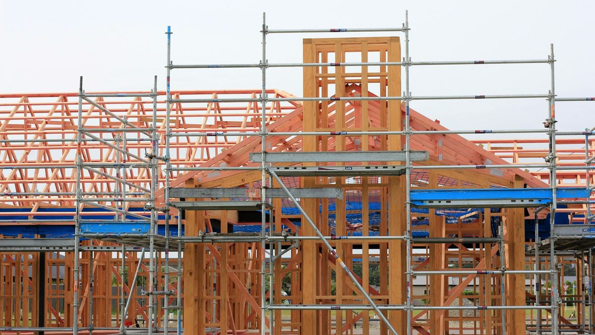 A timber-framed house under construction, its roof trusses exposed behind a scaffold frame.