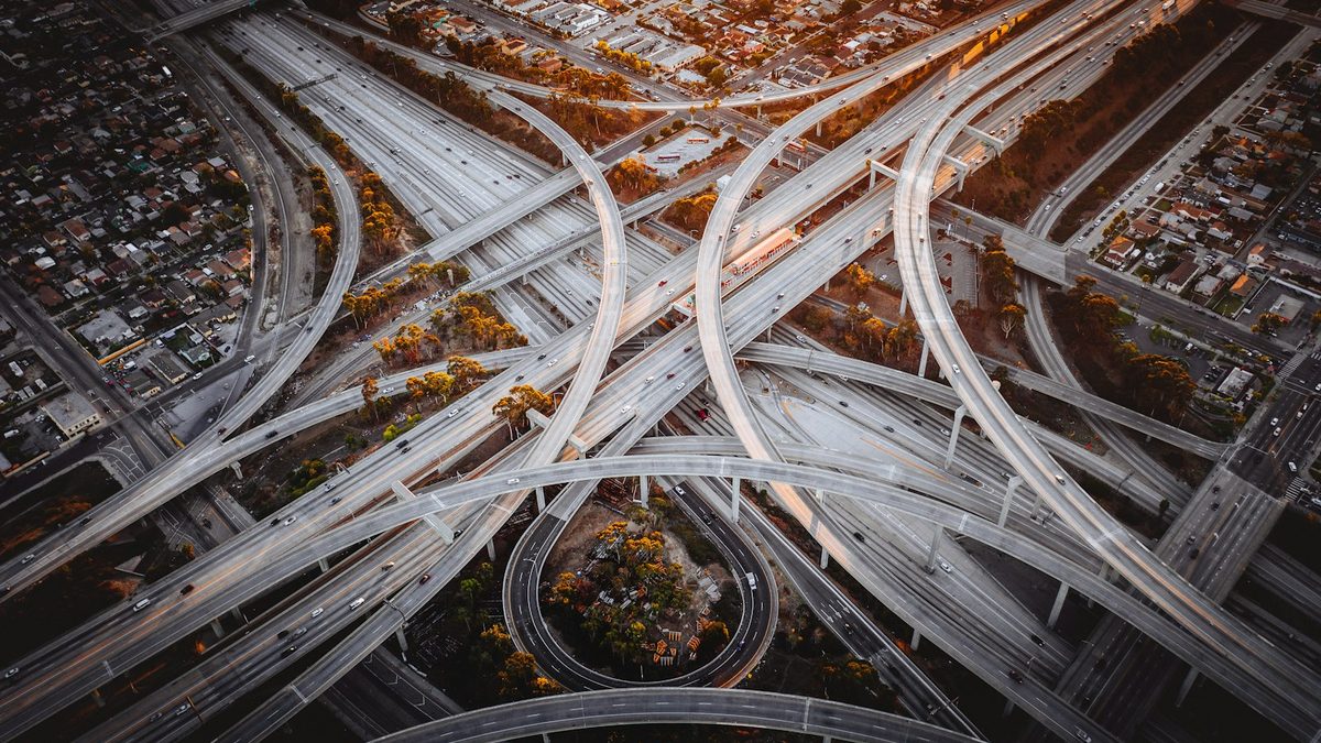 An aerial view of a multi-level freeway interchange at golden hour, curving concrete ramps crossing above straight lanes of traffic, with low-rise housing on either side.