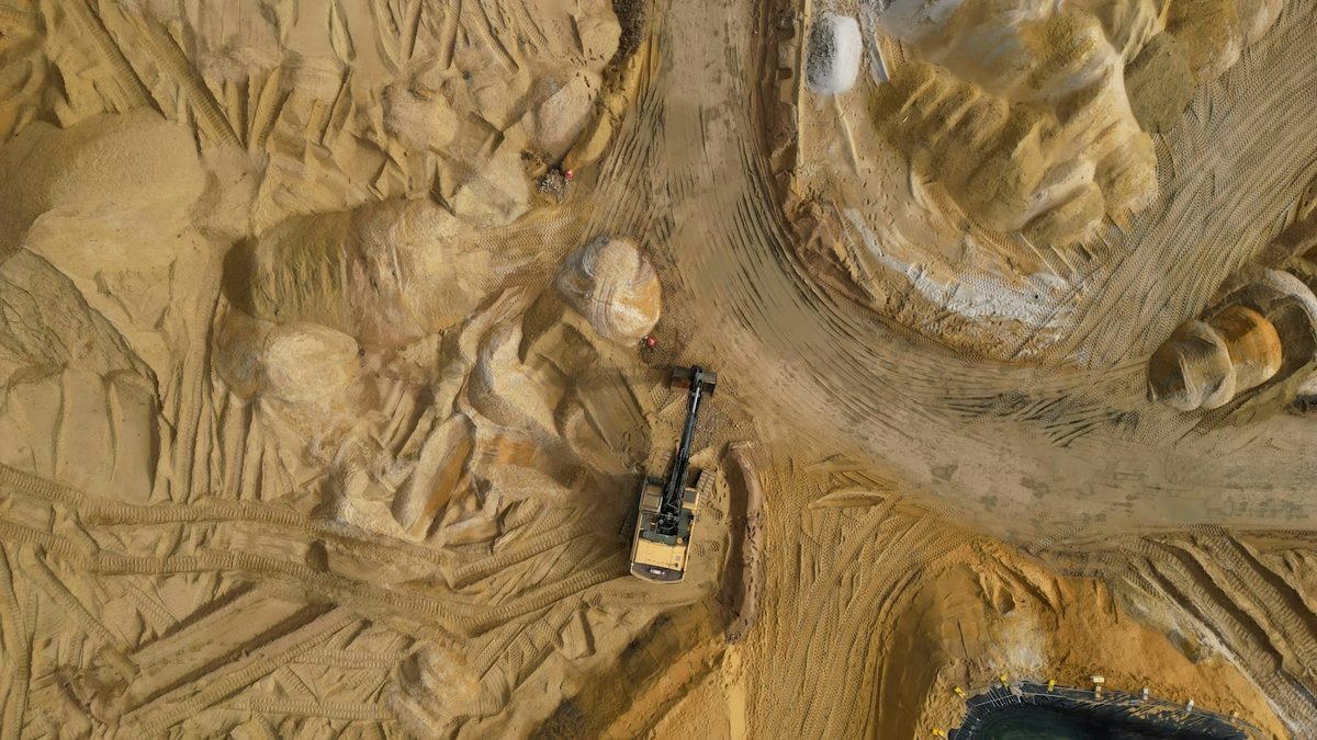 An aerial view of a single excavator working alone on a large sandy earthworks site crossed with machine tracks.