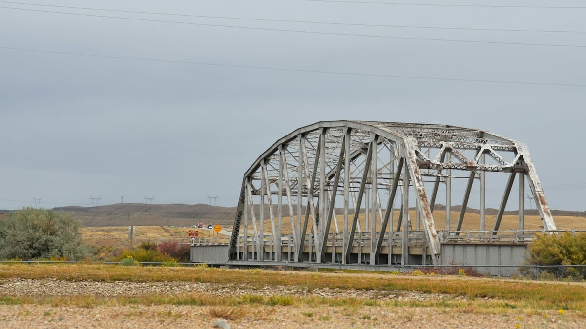 A weathered steel truss road bridge crossing dry open scrubland under an overcast sky, with transmission pylons along the horizon.