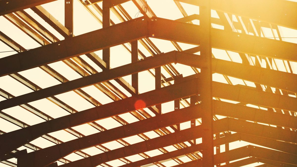 Steel roof beams and columns of an unclad building frame silhouetted against a bright, sunlit sky.