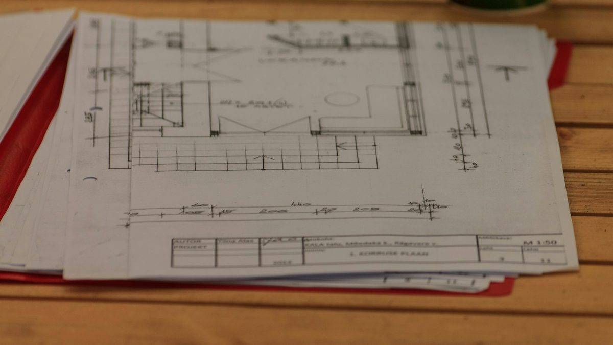 A stack of printed drawing sheets in a red folder on a wooden table, the top sheet showing a floor plan and a title block at 1:50 scale