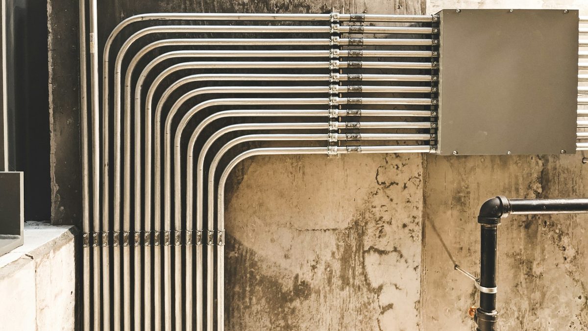 A bank of parallel metal electrical conduits bent in matched right angles across a concrete wall and clamped into a junction box.