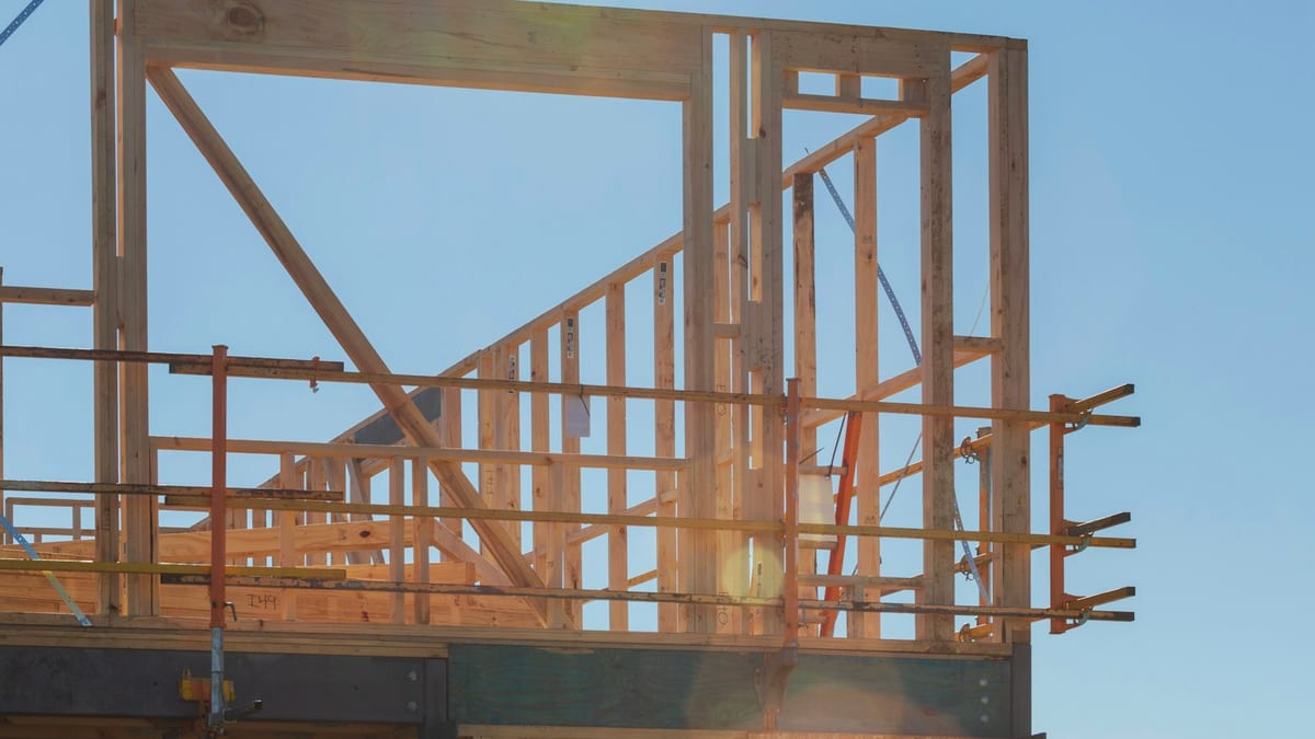 The timber-framed upper storey of a building under construction, with exposed wall studs, a diagonal brace and scaffold guardrails against a clear blue sky.