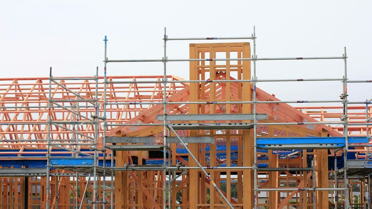 A single-storey timber-framed house under construction on an overcast day, roof trusses exposed and steel scaffolding erected across the front elevation.