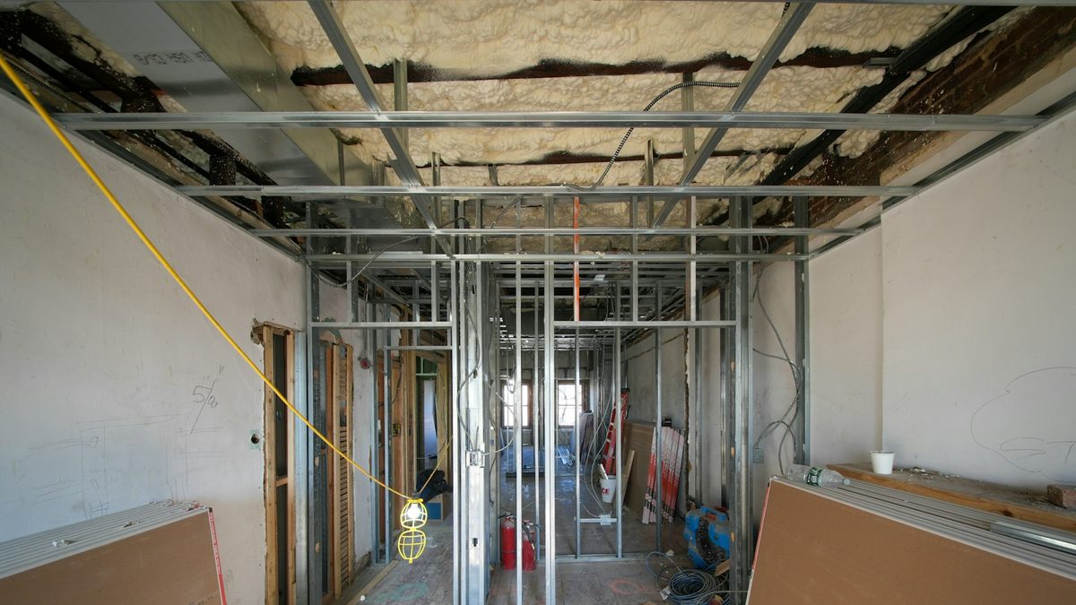 An interior under construction: metal stud partitions and a ceiling grid, exposed spray-foam insulation overhead, and sheets of plasterboard stacked against the walls.