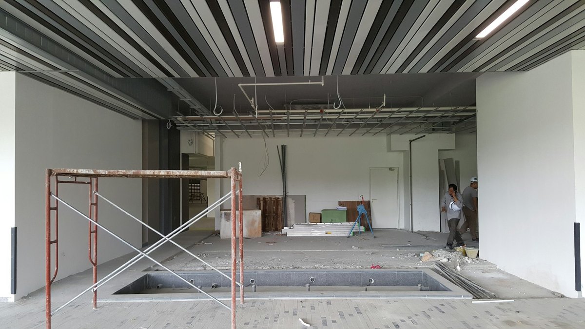 An interior fit-out part-way through: a finished slatted ceiling in the foreground, exposed services and bare ceiling grid beyond, a scaffold frame in the near corner and two workers at the far end of the space.