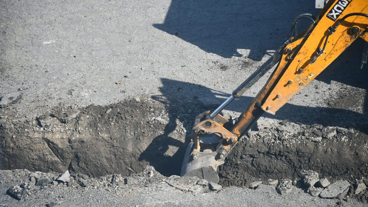 An excavator bucket working in a narrow trench cut through asphalt.