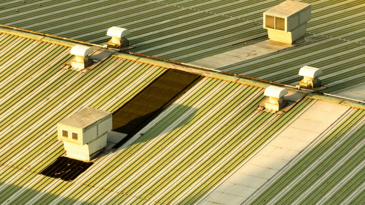 Four ventilation units standing on the green corrugated metal roof of a commercial building, lit by low evening sun.