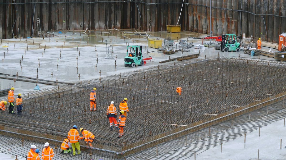 Crews in orange high-visibility gear working across a large steel reinforcement mat in a deep excavation, with forklifts and sheet-piled retaining walls behind them.