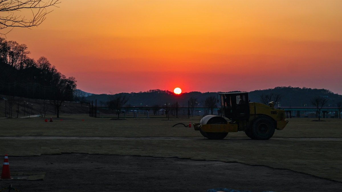 A yellow single-drum soil compactor with an operator in the cab, parked on a levelled earth pad at sunset, traffic cones in the foreground