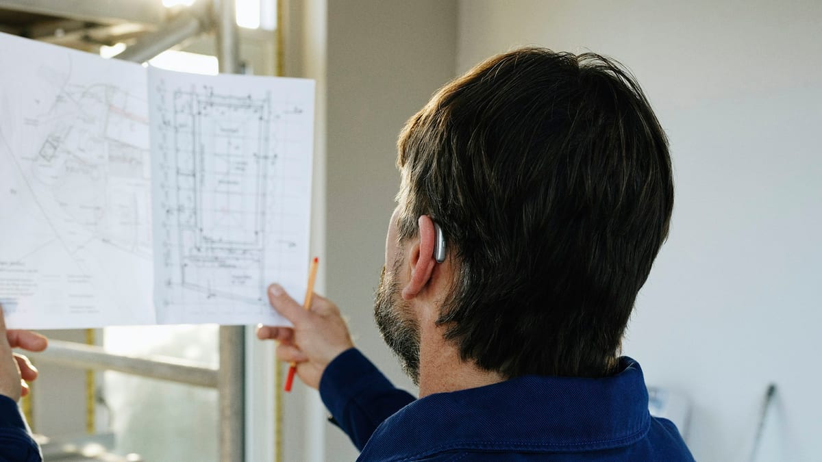 A man in a navy work jacket seen from behind, holding up a pair of printed site plans and a pencil inside an unfinished room with scaffolding by the window