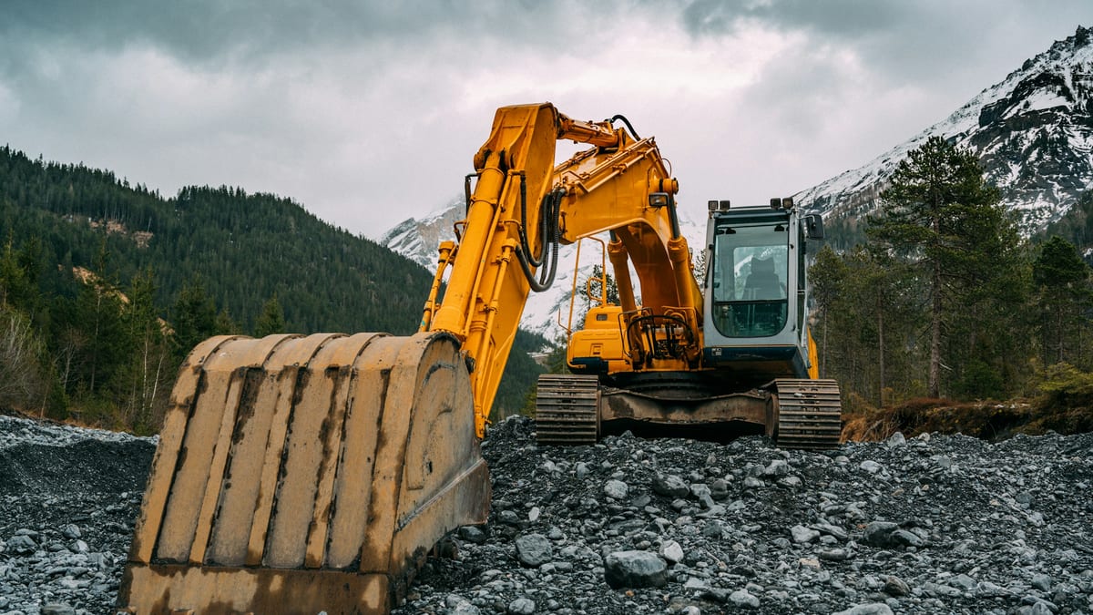 A yellow tracked excavator parked on rocky ground in a mountain valley, bucket resting on the stones.
