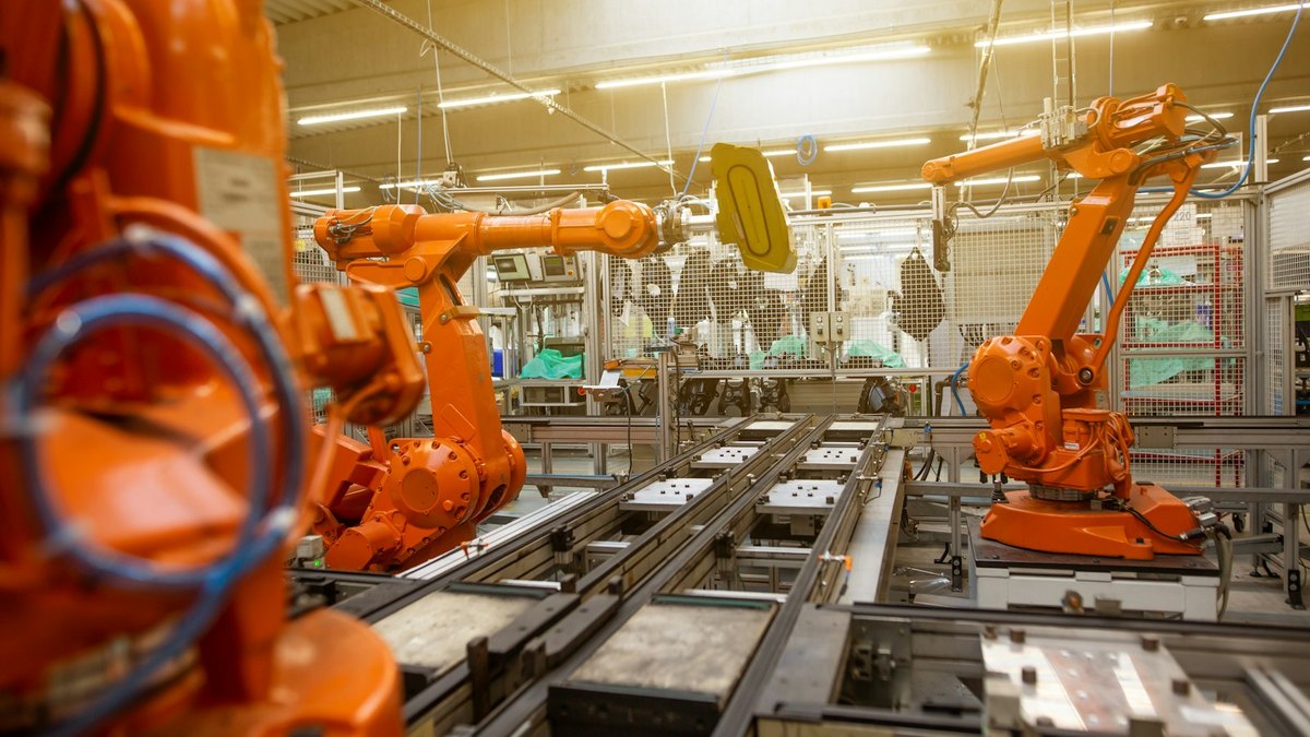 Two orange industrial robot arms either side of a conveyor line inside a factory, with wire-mesh safety guarding behind them