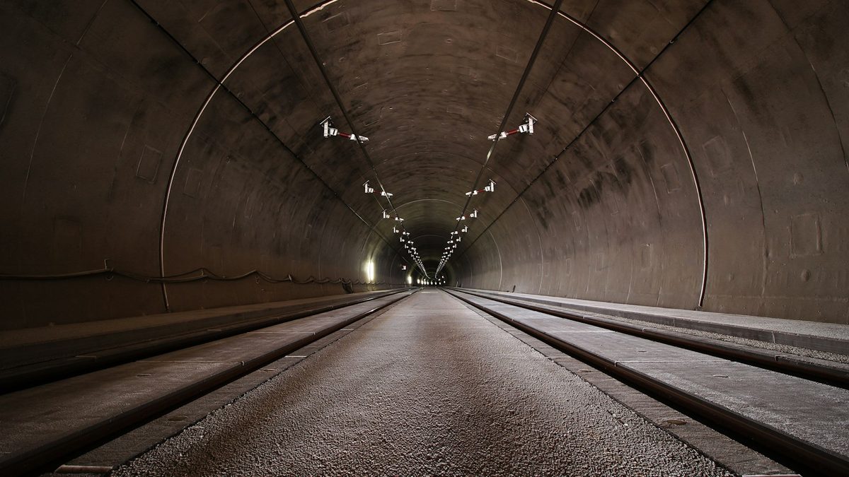 The inside of a finished concrete-lined tunnel, with rail tracks either side of a central strip and lights receding into the distance.
