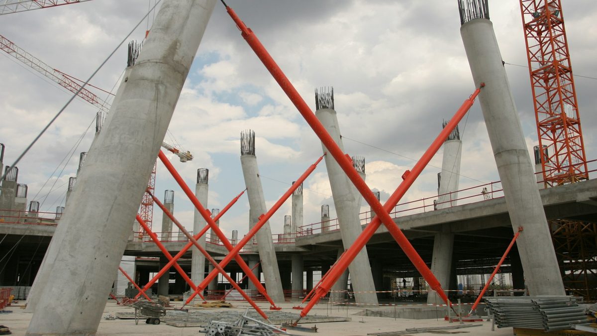 Concrete columns with exposed reinforcement and red raking props on the deck of a large building under construction, with a tower crane behind.