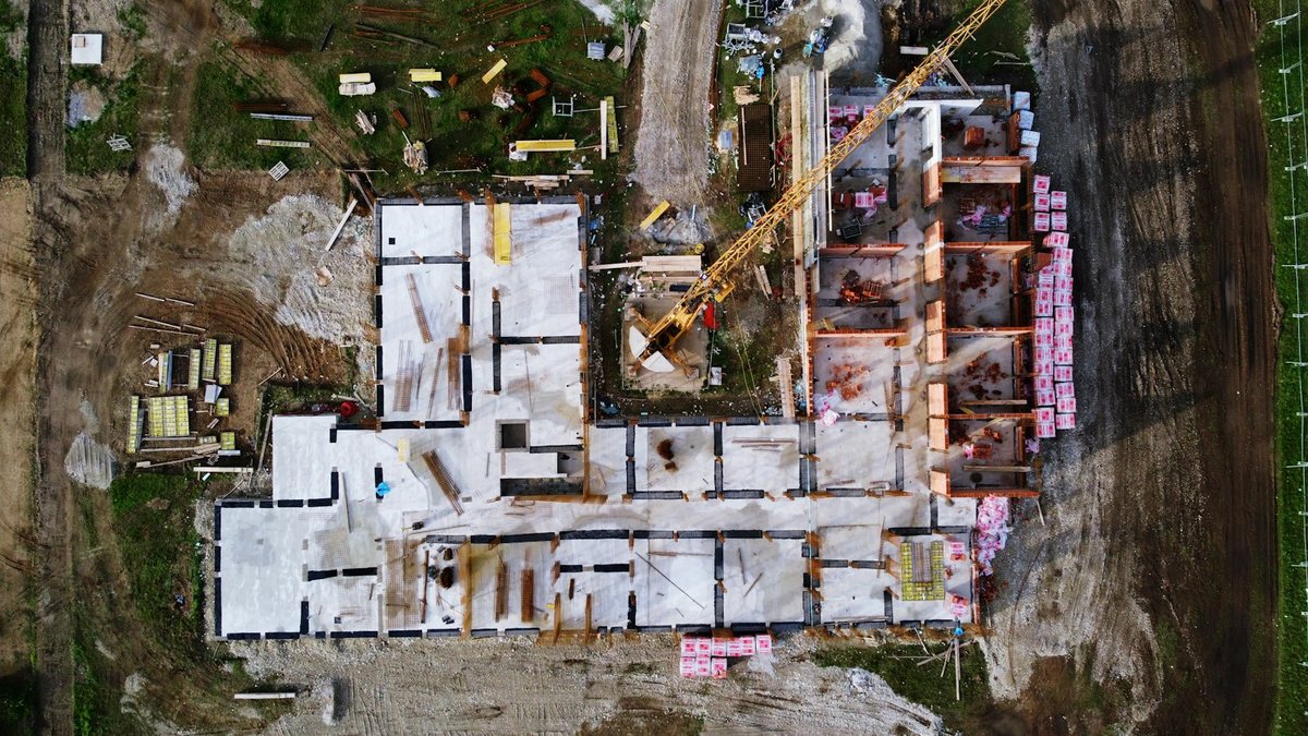 An overhead drone view of a building under construction: pale concrete floor slabs and foundation walls laid out in a plan-like grid, a yellow tower crane angled across the middle, and palletised blocks stacked along one side.