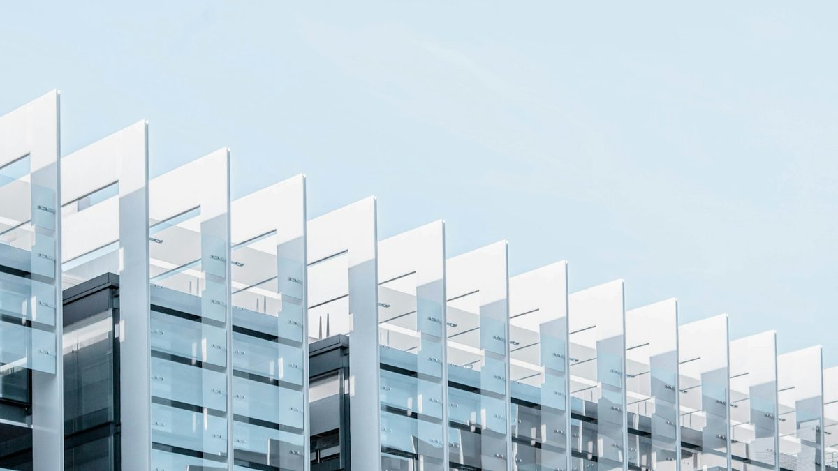 The upper corner of a building clad in a repeating grid of pale glass and metal facade fins, against a clear pale-blue sky.