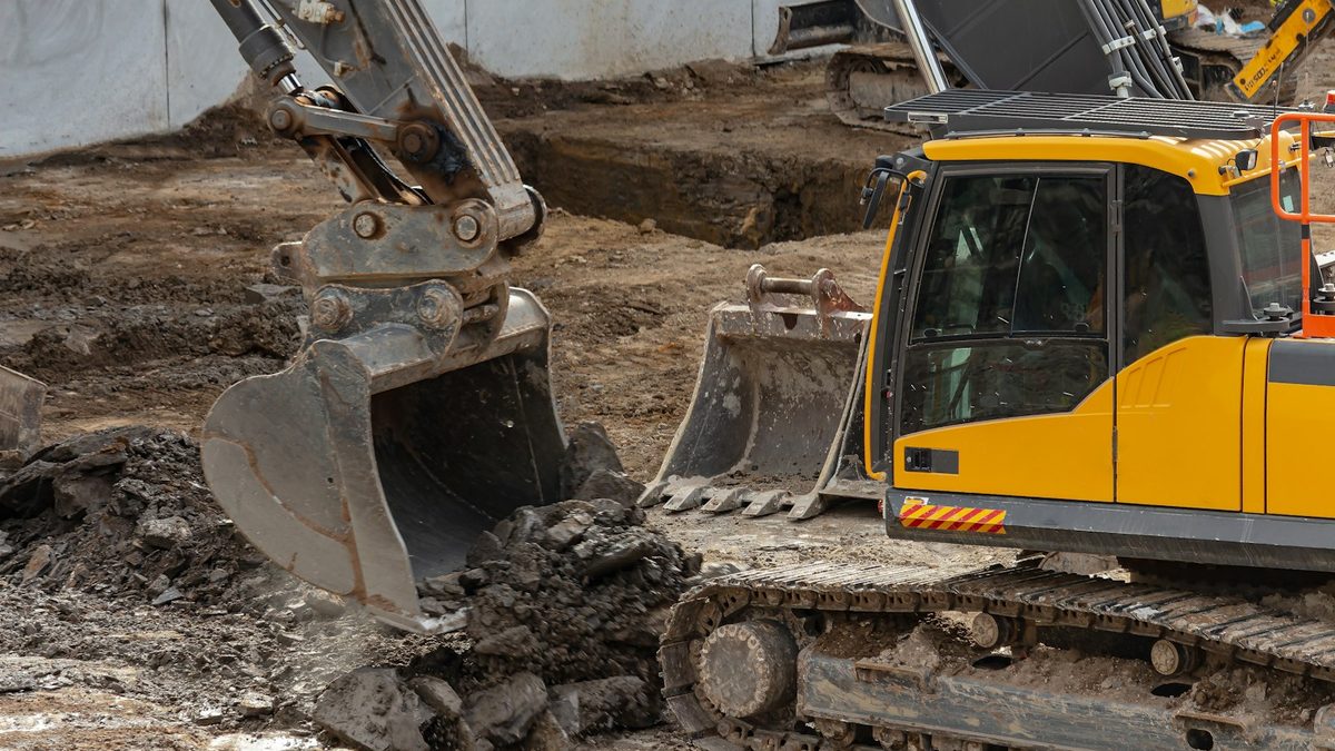 An excavator bucket lowered over broken ground and rubble beside a yellow tracked excavator on an earthworks site.