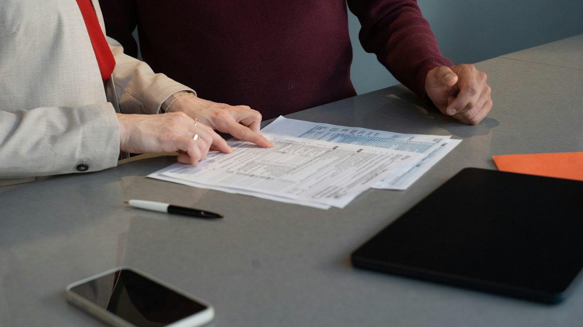 Two people seated at a grey table reviewing printed multi-column forms, one pointing at a line on the page, with a pen, a phone and a tablet on the desk.