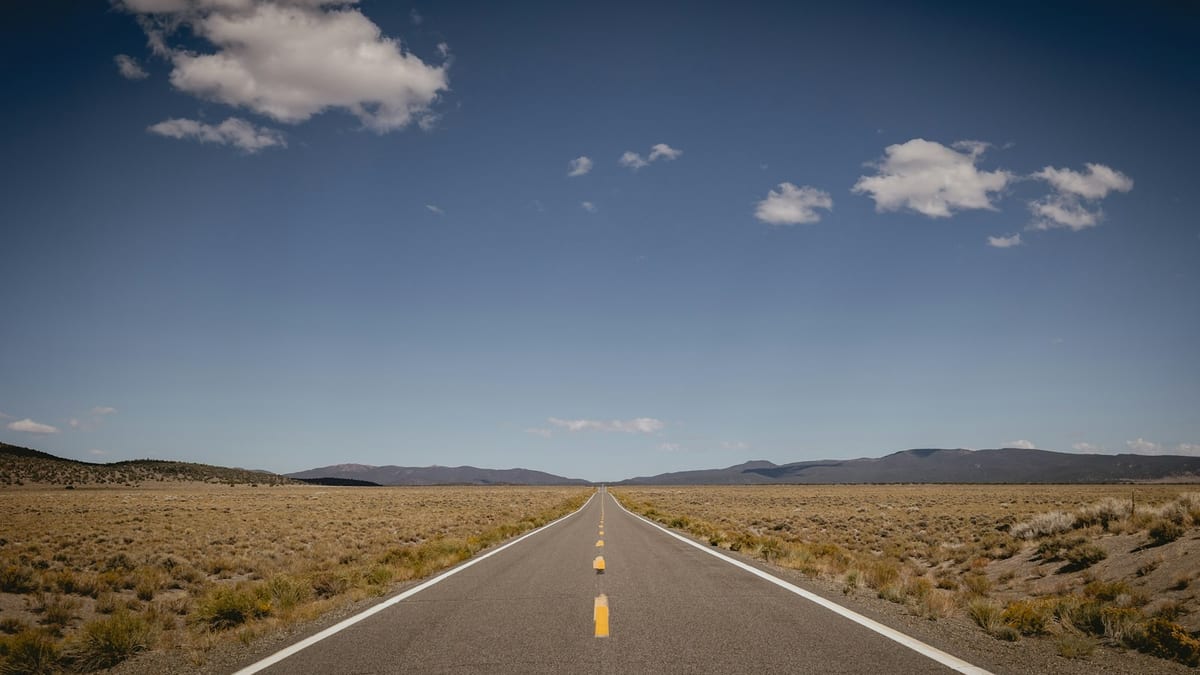 A two-lane highway running straight across open high-desert scrubland towards distant hills, with a yellow centre line and white edge markings.