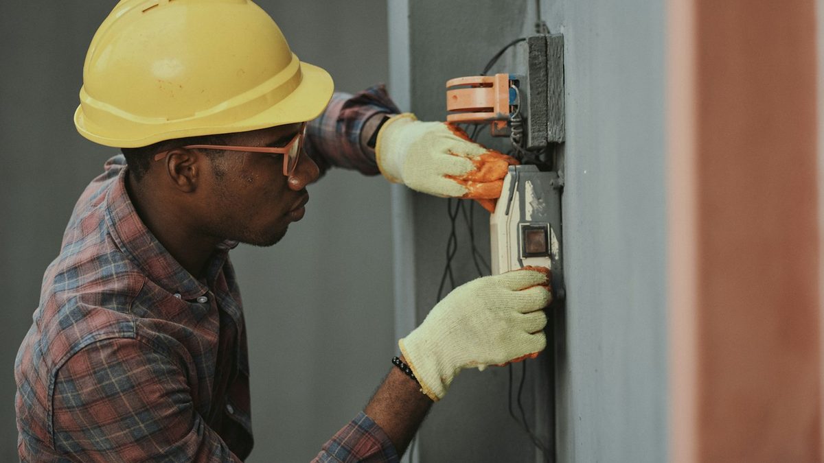 A worker in a yellow hard hat, safety glasses and work gloves wiring an electrical fixture on a grey wall.