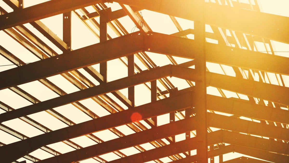 A steel frame under construction seen from below, rows of beams and purlins silhouetted against a bright backlit sky