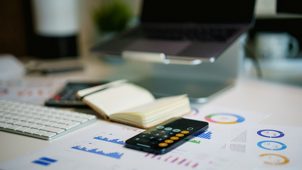 A desk with a laptop, keyboard, calculator, open notebook and printed financial charts.