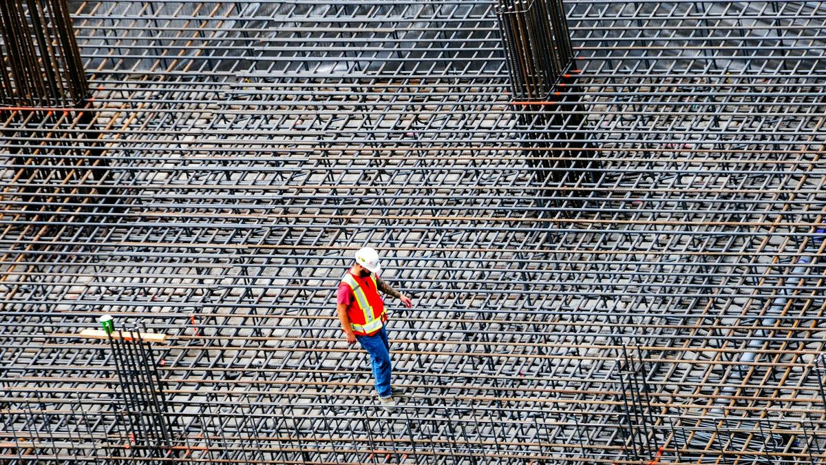A lone worker in a hard hat and high-visibility vest walking across a large rebar mat on a construction site.