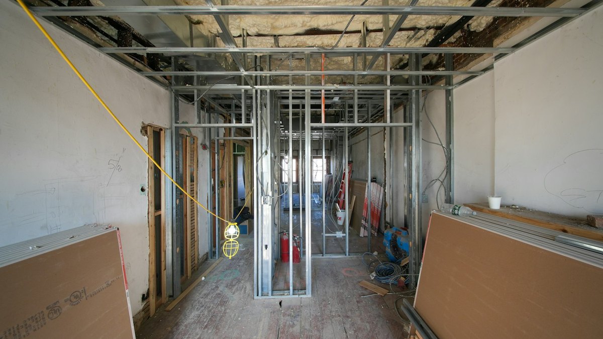 An interior room under construction, with metal stud partitions, an exposed ceiling and stacked drywall panels in the foreground