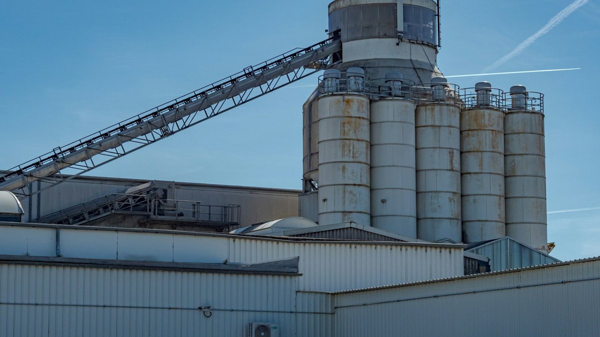 A cement plant: a cluster of weathered steel silos and a long elevated conveyor rising above corrugated sheds, against a clear blue sky.