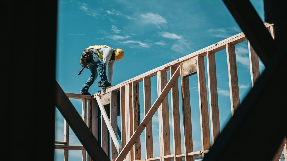 A construction worker in a yellow hard hat and high-visibility vest bends over the top plate of a timber wall frame, seen from below against a blue sky