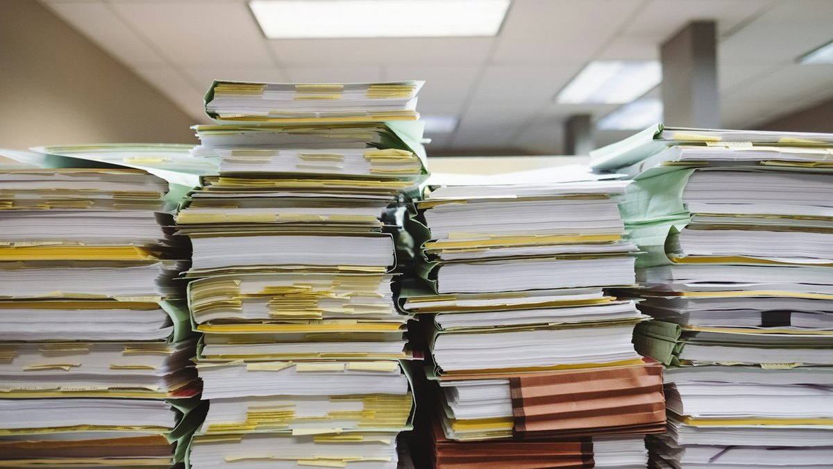 Tall stacks of paper files and folders, marked with coloured index tabs, piled up in an office.