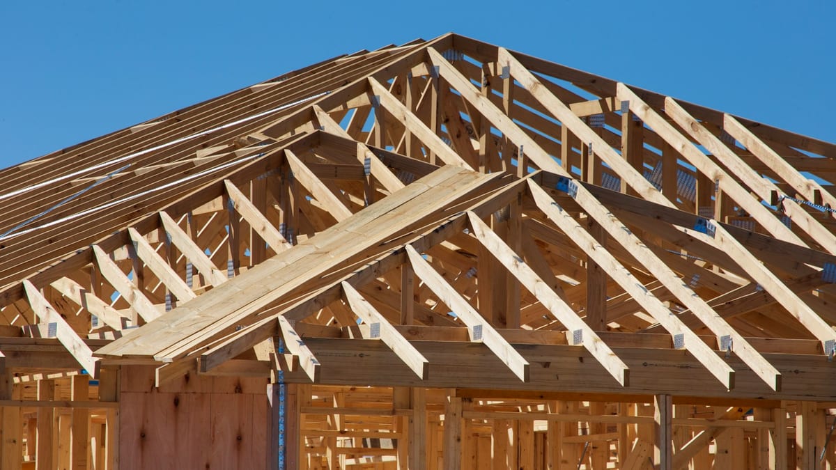 Timber roof trusses and stud framing of a house under construction against a clear blue sky.