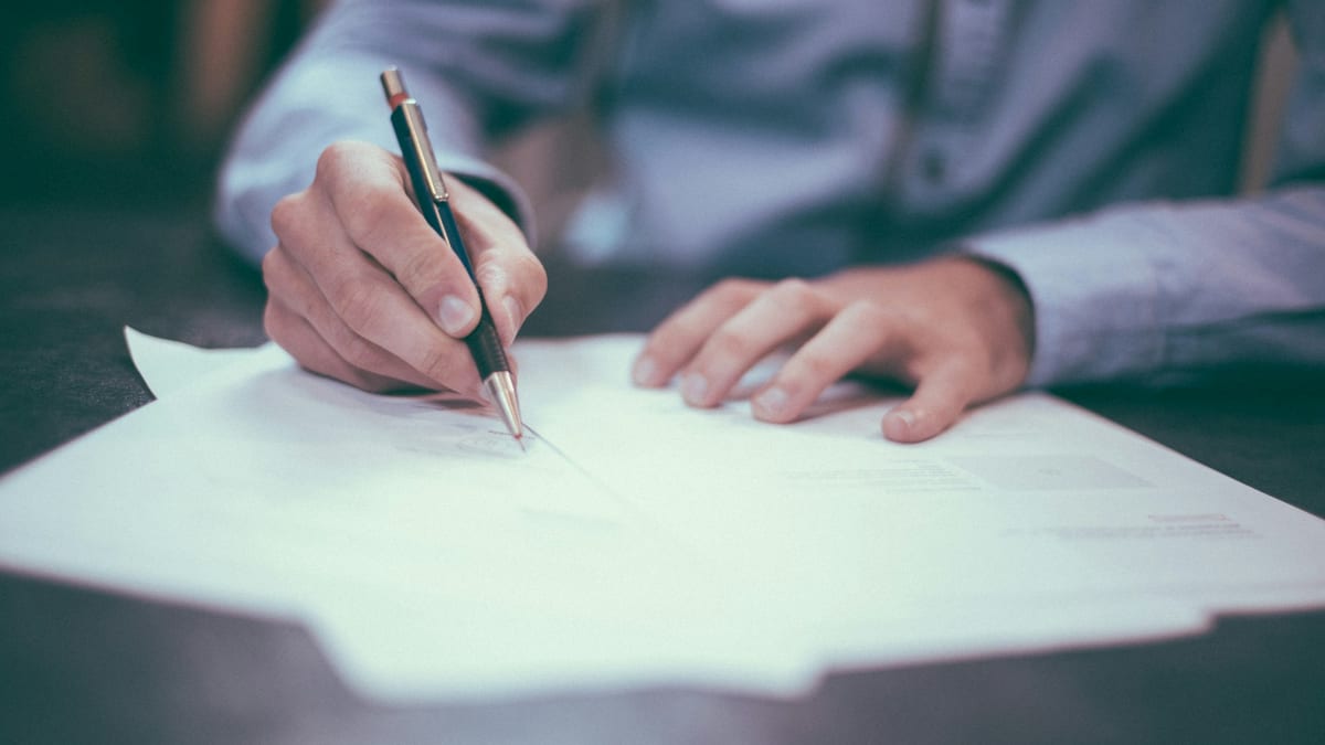 Close-up of a person's hands and a pen resting on loose sheets of paper on a dark desk, as if about to sign a document.