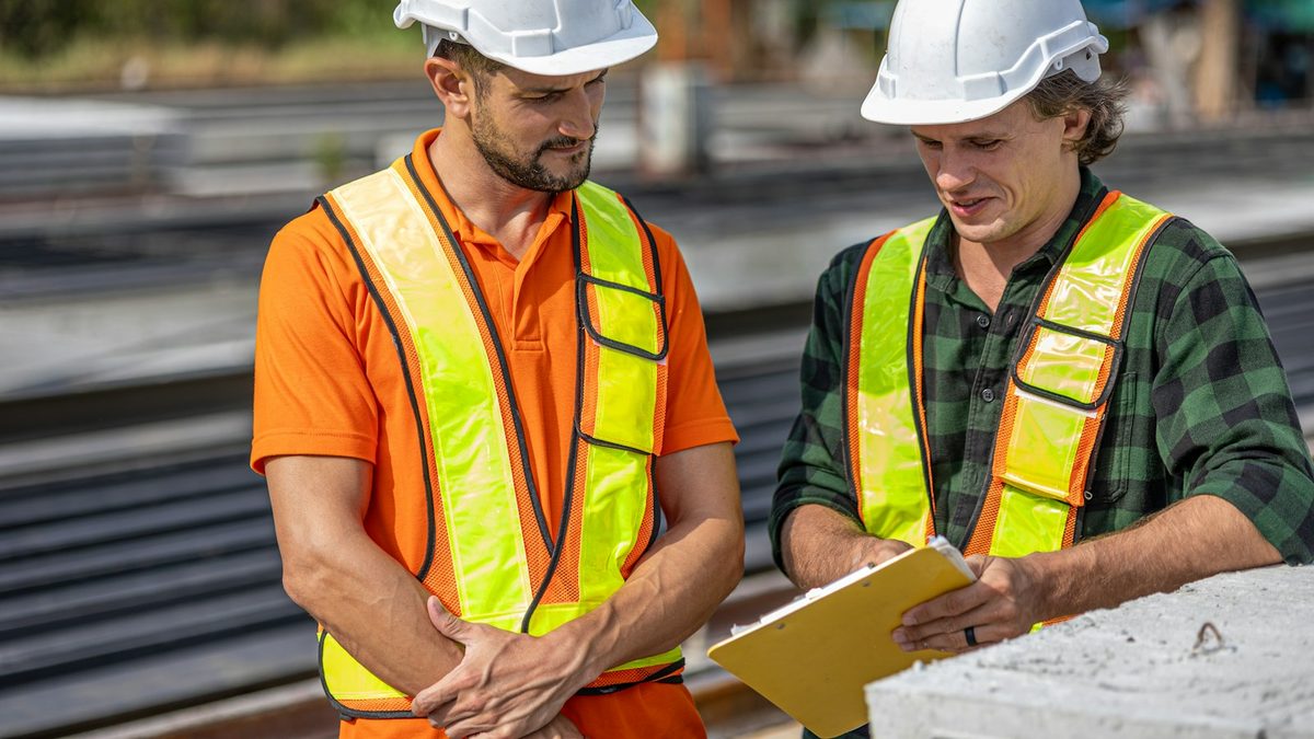 Two construction workers in hard hats and hi-vis vests reviewing a document on a clipboard.