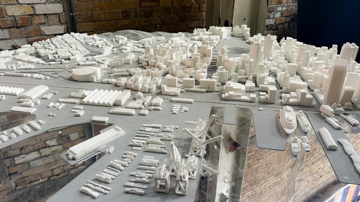 A white architectural massing model of a city and its harbour, with dock cranes and ships, on a grey baseboard in a brick-walled room