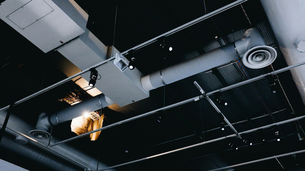 Exposed ventilation ductwork with circular diffusers running beneath a dark commercial ceiling, alongside lighting track and cable containment