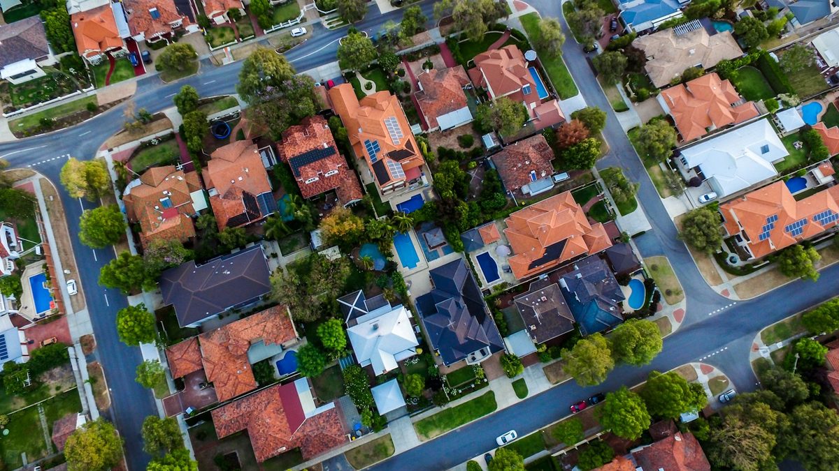 A straight-down aerial view of a suburban neighbourhood: tiled pitched roofs, several fitted with solar panels, with swimming pools, trees and curving streets between them.