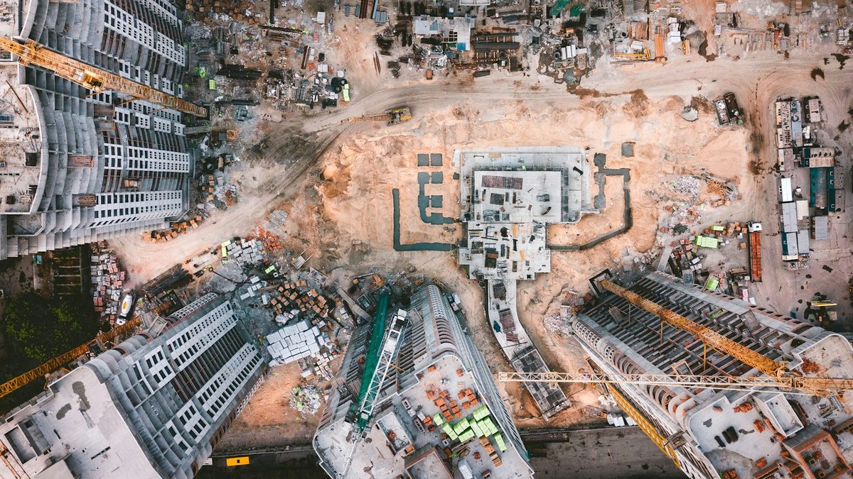 An overhead aerial view of a large residential construction site, with several apartment blocks under construction around a cleared central plot, tower cranes and stacked materials.