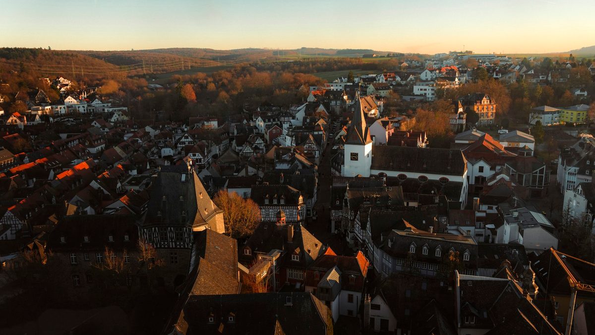 An elevated view over the rooftops of a German town at sunset, with half-timbered houses, a church tower and wooded hills beyond.