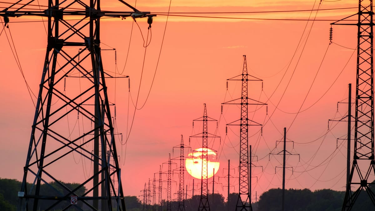 Rows of high-voltage electricity transmission pylons silhouetted against an orange sunset.