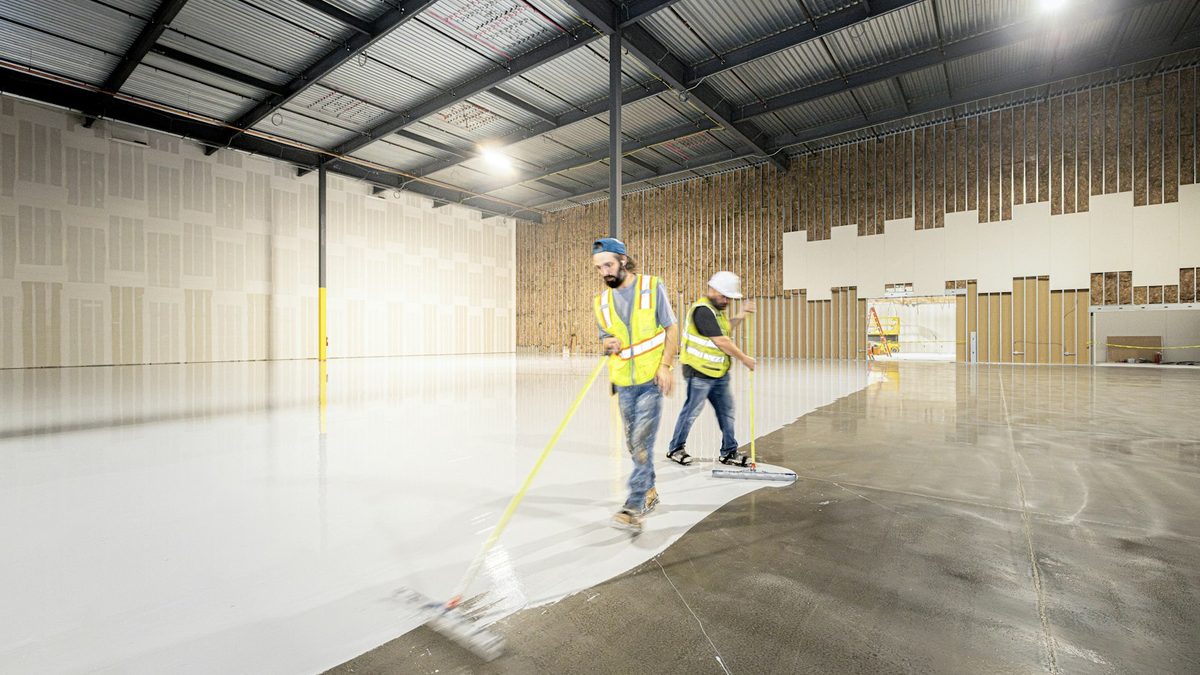Two workers in high-visibility vests spreading a coating across the floor of a large empty commercial building