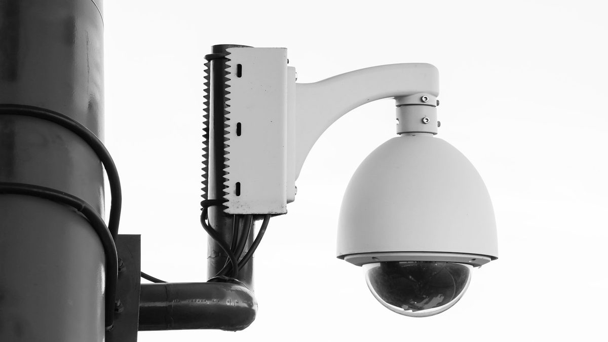 A white dome security camera on a curved bracket mounted to a dark metal pole, photographed from below against a blank white sky.