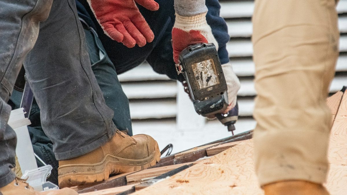 A gloved hand holding a cordless impact driver, driving a screw into timber roof decking, with another worker's legs and boots in the foreground.
